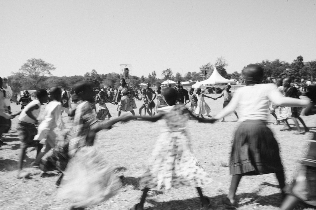 Children and adults form a circle, holding hands and dancing in an open field at an outdoor festival in daylight, black-and-white.